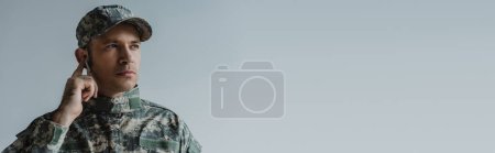 soldier in uniform and cap adjusting wireless earphones isolated on grey, banner 