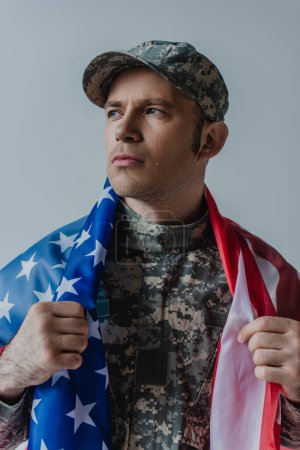trauriger amerikanischer Soldat mit Flagge der Vereinigten Staaten, während er am Gedenktag weint, isoliert auf grau 