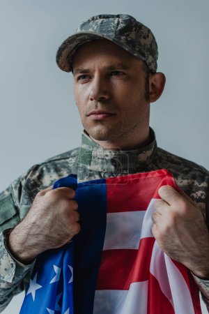 Verärgerter amerikanischer Soldat hält Flagge der Vereinigten Staaten, während er am Gedenktag weint, isoliert auf grau 