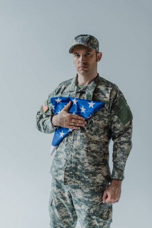 gut aussehender Soldat in Militäruniform mit gefalteter Flagge der Vereinigten Staaten am Gedenktag isoliert auf grau 