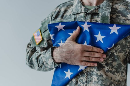 Teilansicht eines amerikanischen Soldaten mit gefalteter Flagge der Vereinigten Staaten am Gedenktag isoliert auf grau 