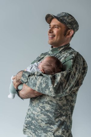 fröhlicher Vater in Militäruniform und Mütze umarmt neugeborenen Jungen isoliert auf grau 