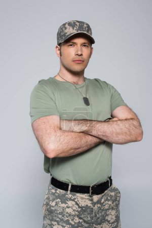 soldado en camiseta y gorra de pie con los brazos cruzados durante el día conmemorativo aislado en gris 