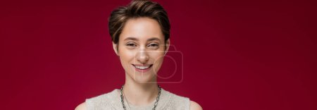 Photo for Cheerful young woman with short hair smiling and looking at camera on dark red background, banner - Royalty Free Image