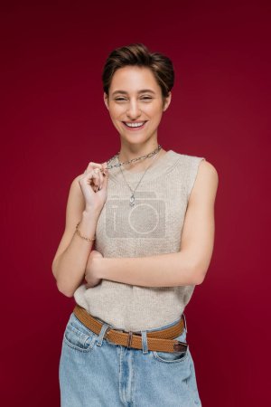 pretty young woman with short hair smiling and pulling necklace chain on dark red background