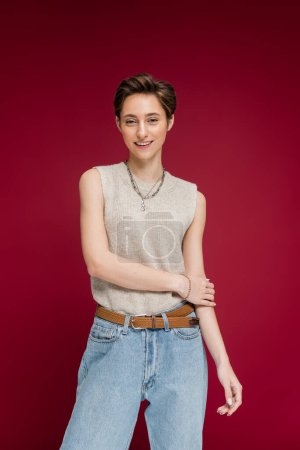 overjoyed young woman with short hair posing in jeans and sleeveless shirt on dark red background
