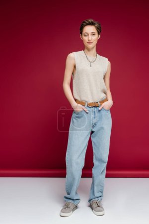 full length of pleased young woman with short hair posing with hands in pockets of jeans on dark red background