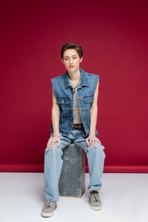 full length of stylish young woman with short hair sitting in denim outfit on dark red background