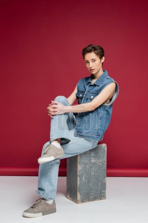 full length of pretty young model in denim outfit sitting on dark red background