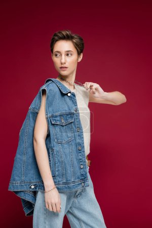 Photo for Pretty young model in denim vest pulling necklace chain while posing on dark red background - Royalty Free Image