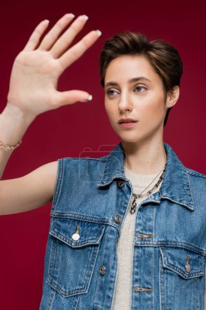 young model in denim vest standing with outstretched hand on maroon background