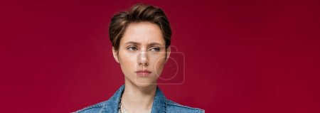 suspicious young woman with short hair looking away on burgundy background, banner 