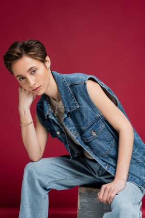stylish model in denim outfit with vest sitting on burgundy background 