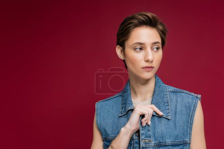 stylish model in denim outfit with vest looking away on burgundy background 