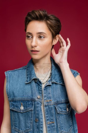stylish model in denim outfit with vest fixing her short hair on burgundy background 