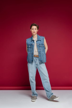 full length of stylish model in denim outfit with vest standing on burgundy background 