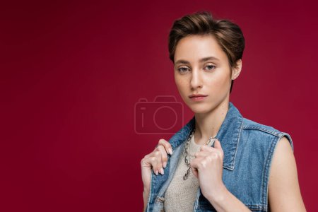 stylish model in adjusting denim vest while posing on burgundy background 