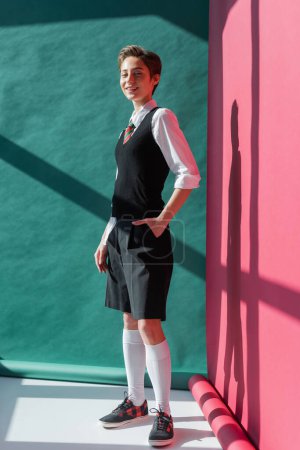 Photo for Full length of happy young student with short hair posing with hand in pocket of black shorts on pink and green - Royalty Free Image