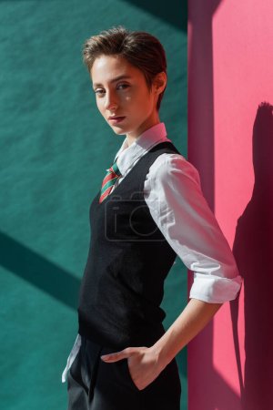 Photo for Stylish young student with short hair posing with hand in pocket of black shorts on pink and green - Royalty Free Image