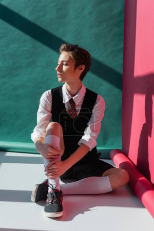 full length of stylish young student with short hair sitting in school uniform on pink and green 
