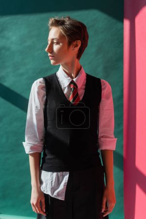 stylish student with short hair standing in school uniform on pink and green 