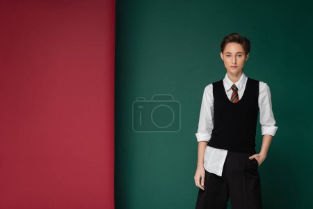 stylish young student with short hair posing with hand in pocket on green and maroon background 