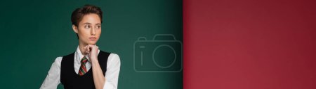 stylish young student with short hair posing in school uniform and looking away on green and maroon background, banner 