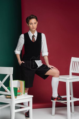 Photo for Stylish young woman with short hair posing in school uniform around chairs and books on green and pink background - Royalty Free Image