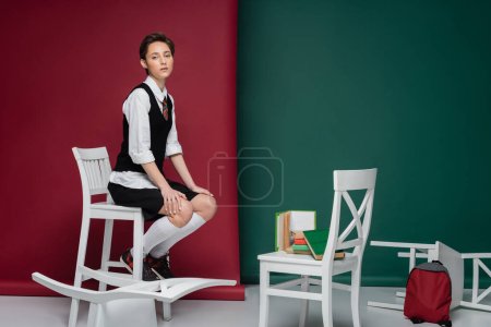 volle Länge der stilvollen jungen Frau mit kurzen Haaren sitzt auf einem Stuhl in der Nähe von Büchern auf grünem und rosa Hintergrund 