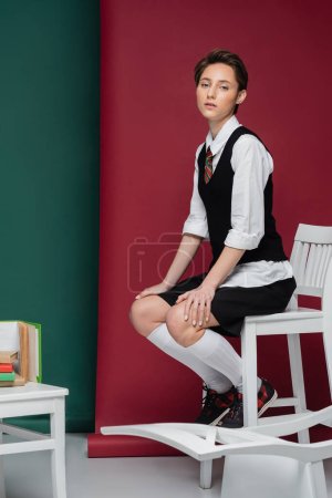 volle Länge der stilvollen junge Student mit kurzen Haaren sitzt auf Stuhl in der Nähe von Büchern auf grünem und rotem Hintergrund 