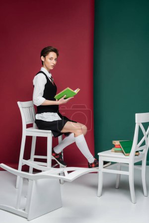 Foto de Longitud completa de la mujer joven con estilo con el pelo corto sentado en la silla y libro de lectura sobre fondo verde y rojo - Imagen libre de derechos
