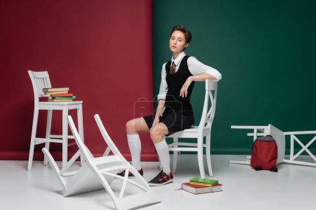 volle Länge der stilvollen jungen Frau mit kurzen Haaren sitzt auf weißem Stuhl zwischen Büchern auf grünem und rotem Hintergrund 