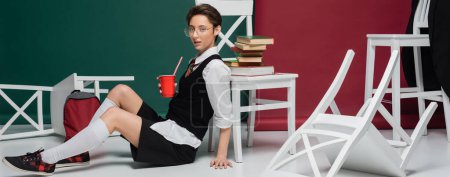 stylish student in eyeglasses holding drink in plastic cup near chairs and books on green and burgundy background, banner 