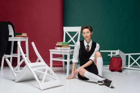 full length of stylish young student in eyeglasses sitting around chairs with books on green and pink background 