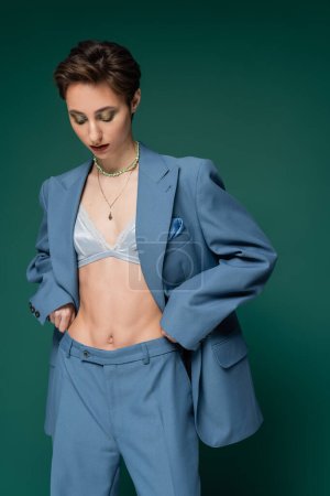 young woman with short hair posing in satin bra and blue pantsuit on turquoise green background 