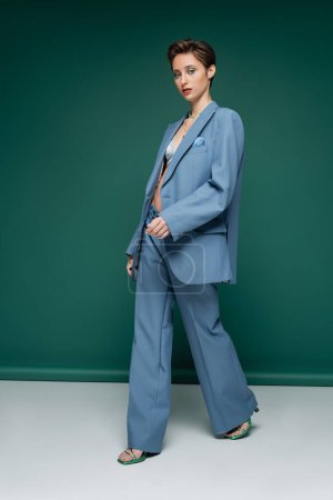 full length of young woman with short hair walking in blue pantsuit with bra on turquoise green background 