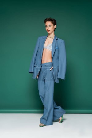 full length of young woman with short hair posing in blue pantsuit on turquoise green background 