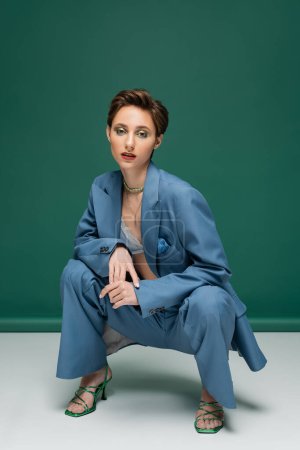 young woman with short hair sitting on haunches while posing in trendy suit and heeled sandals on turquoise white background 