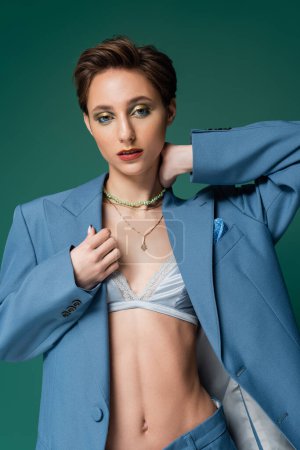 young model with short hair posing in blue jacket with silk bra underneath on turquoise background 