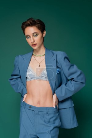 young woman in blue suit with silk bra underneath posing with hands on hips on turquoise green background 