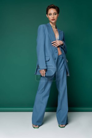 full length of young model with short hair posing in blue suit on turquoise white background 