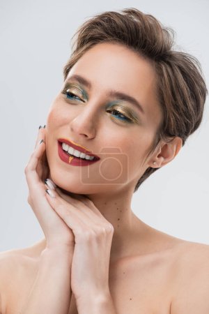 glückliche junge Frau mit kurzen Haaren und hellem Make-up, die isoliert von grau wegschaut 