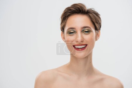 Foto de Mujer joven llena de alegría con el pelo corto y maquillaje brillante mirando a la cámara aislada en gris - Imagen libre de derechos