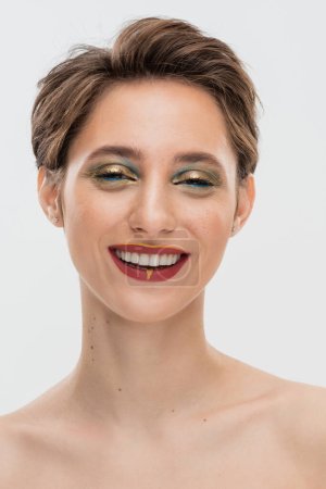 Foto de Mujer joven positiva con pelo corto y maquillaje brillante mirando a la cámara aislada en gris - Imagen libre de derechos