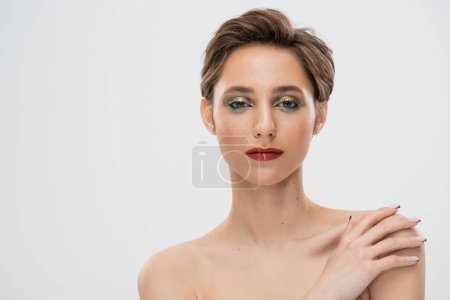 Foto de Retrato de mujer joven con maquillaje brillante tocando hombro desnudo y mirando a la cámara aislada en gris - Imagen libre de derechos