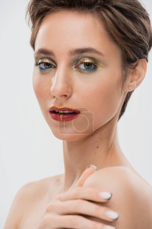 Porträt eines jungen Models mit hellem Make-up, das die nackte Schulter berührt und isoliert auf grau in die Kamera blickt 