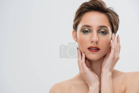 portrait de jeune modèle avec un maquillage lumineux touchant les joues et regardant la caméra isolée sur gris 