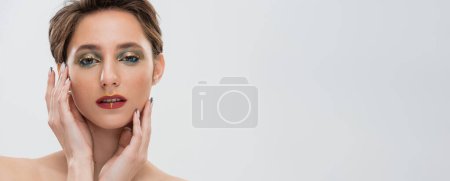 portrait of young woman with bright makeup touching cheeks and looking at camera isolated on grey, banner 