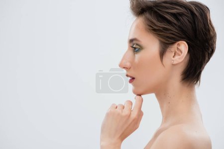 Téléchargez les photos : Vue latérale de la jeune femme avec un maquillage lumineux touchant menton isolé sur gris - en image libre de droit