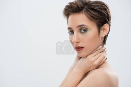 retrato de mujer joven con maquillaje brillante tocando el cuello y mirando a la cámara aislada en gris